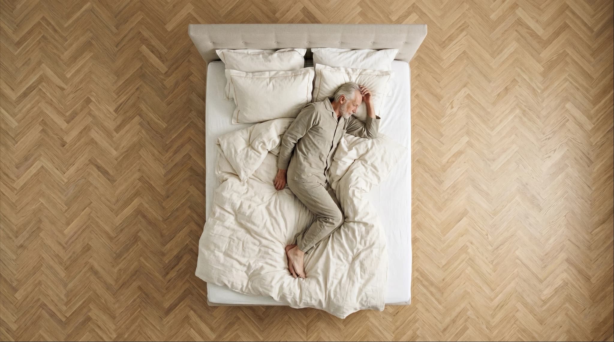 Overhead view of a man in linen pyjamas asleep on a Mattsons bed with cream bedding and a tufted headboard, on a herringbone parquet floor